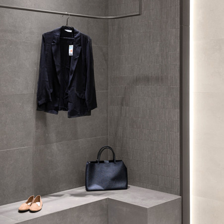 Een zwarte blazer hangt aan een haak in een minimalistische kleedkamer met donkergrijze tegels, naast een zwarte handtas en beig