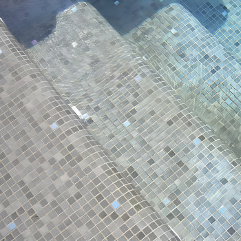 Glasfliesen bilden in Wasser getauchte Stufen und erzeugen ein schimmerndes Muster unter klarem, sanft fließendem Poolwasser.