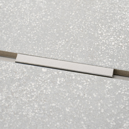 Ein rechteckiger, silberner Einsatz durchbricht das einheitliche Muster der hellgrauen, gesprenkelten Fliesen und bildet einen s