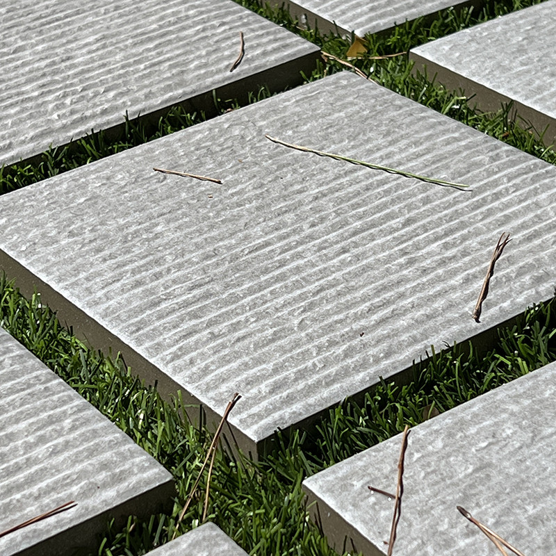 Square stone tiles lie evenly spaced on lush green grass, featuring textured ridges with scattered slender twigs resting on thei