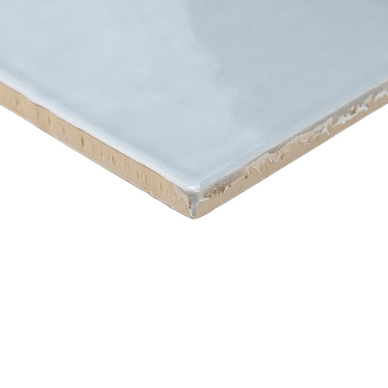 Eine glänzende, hellblaue Keramikfliese mit glatter Oberfläche liegt schräg und zeigt ihre unbearbeitete beige Kante vor einem w