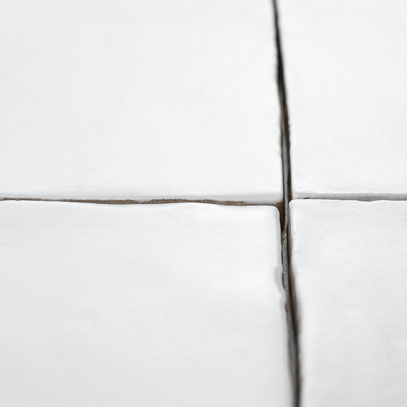 White ceramic tiles form a grid with visible grout lines in a clean, minimalistic space. Edges slightly uneven, creating subtle 