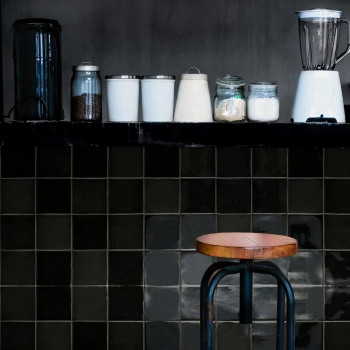 Op een aanrecht staan ​​een blender, potten en een jerrycan. Beneden staat een houten kruk tegen donker betegelde muren.