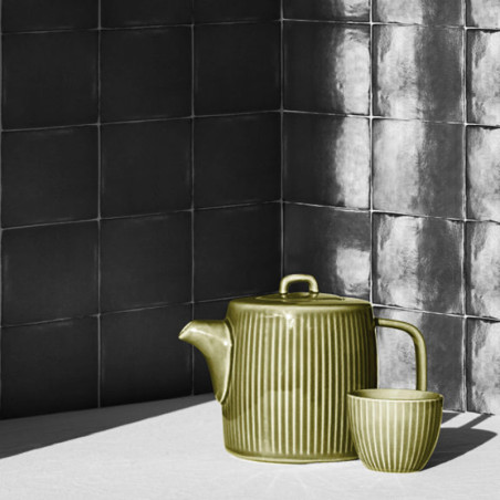 Une théière verte nervurée et une tasse assortie sont exposées au soleil sur un fond de carreaux carrés sombres et texturés.