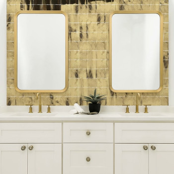 Dois espelhos retangulares com molduras douradas refletem contra uma parede texturizada com azulejos dourados. Abaixo, pias dupl