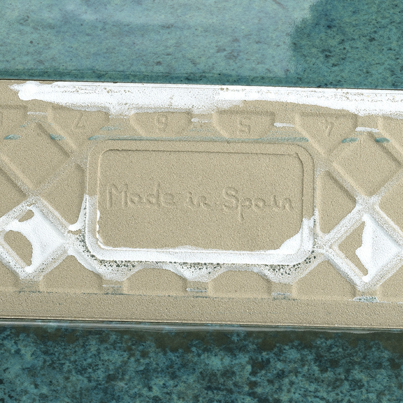 Die rechteckige Fliese mit der Aufschrift „Made in Spain“ liegt flach, teilweise mit Sand bedeckt, auf einer strukturierten grün