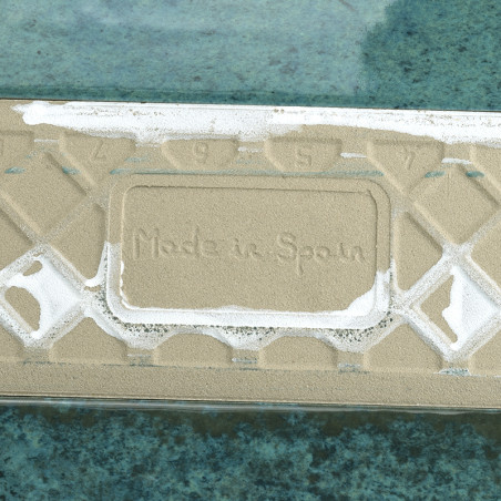 Le carreau rectangulaire, portant l'inscription « Made in Spain », repose à plat, partiellement recouvert de sable, sur une surf