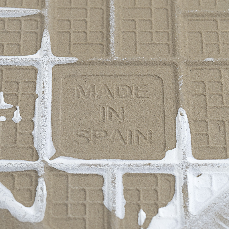 Un carreau texturé affiche les mots « MADE IN SPAIN » au milieu du coulis séché, disposés selon un motif en grille.