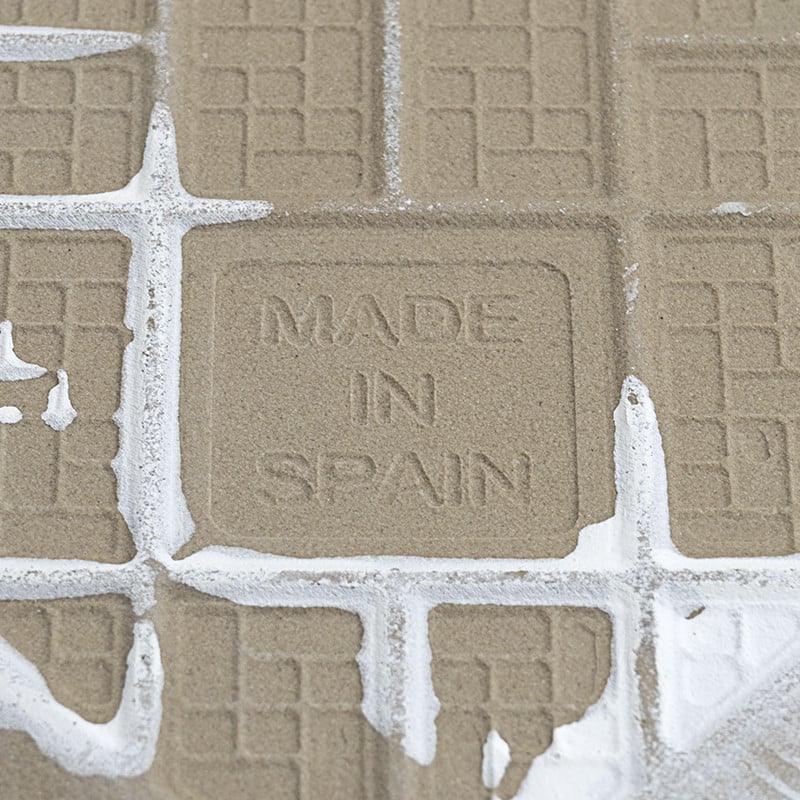 Square tile displays `MADE IN SPAIN` embossed, surrounded by grout residue. The background consists of similar grid-patterned ti