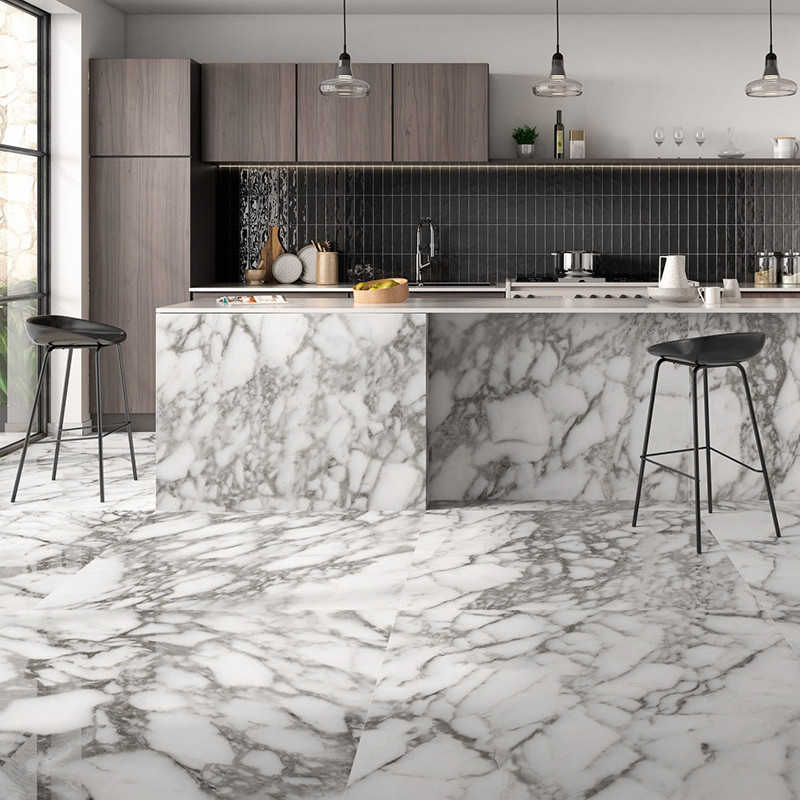 Azulejo tipo mármol blanco con vetas grises usado en suelo e isla; cocina moderna con armarios de madera oscura, salpicadero negro, taburetes altos y lámparas colgantes.