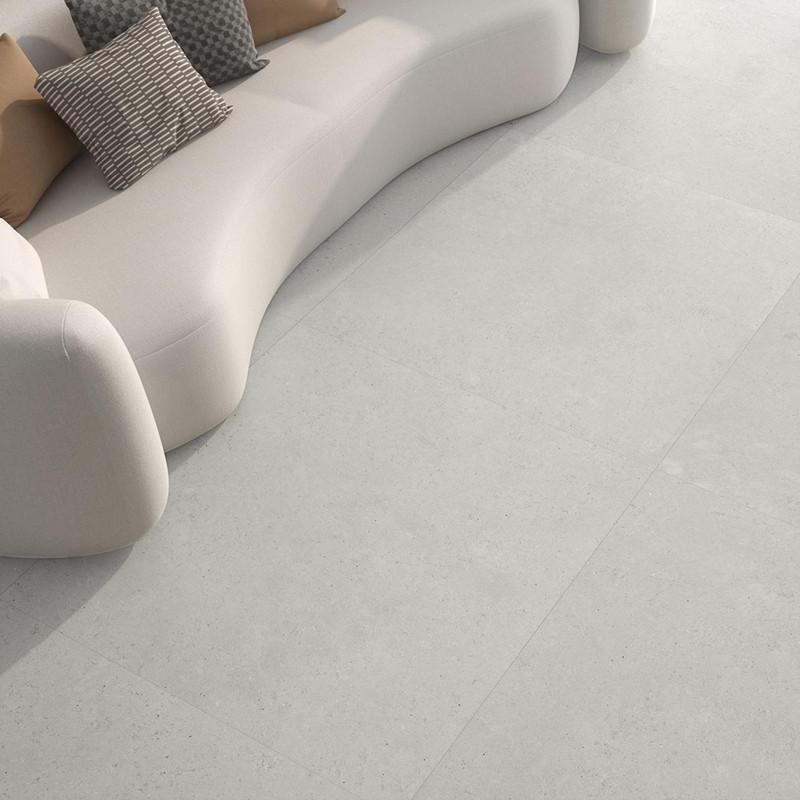Azulejo gris claro mate en suelo amplio junto a sofá curvo beige con varios cojines; juntas rectilíneas y textura sutil tipo piedra.