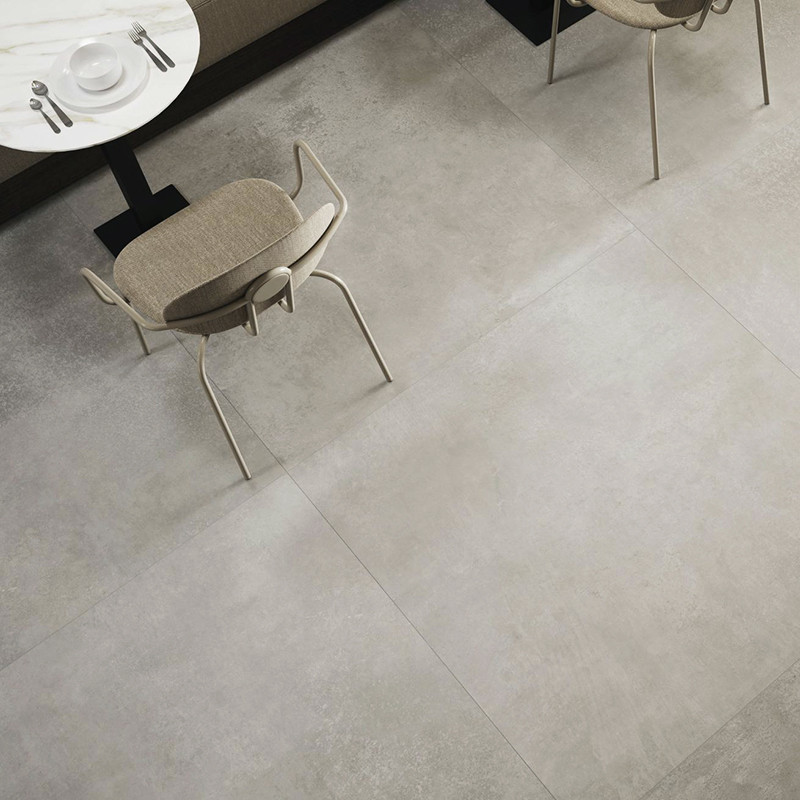 Carrelage grand format gris clair mat au premier plan; contexte: table ronde noire dressée et deux chaises beiges, coin de café/restaurant.
