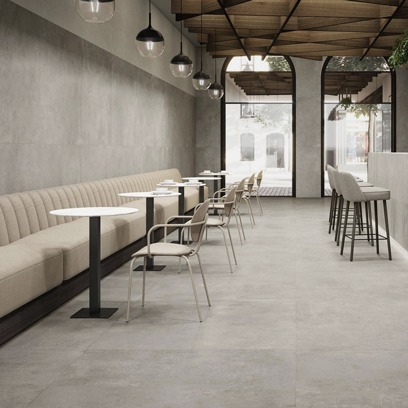 Azulejo gris claro mate, formato grande; en cafetería minimalista con banqueta beige, mesas y sillas blancas, lámparas colgantes, barra alta y arcos que abren al exterior.