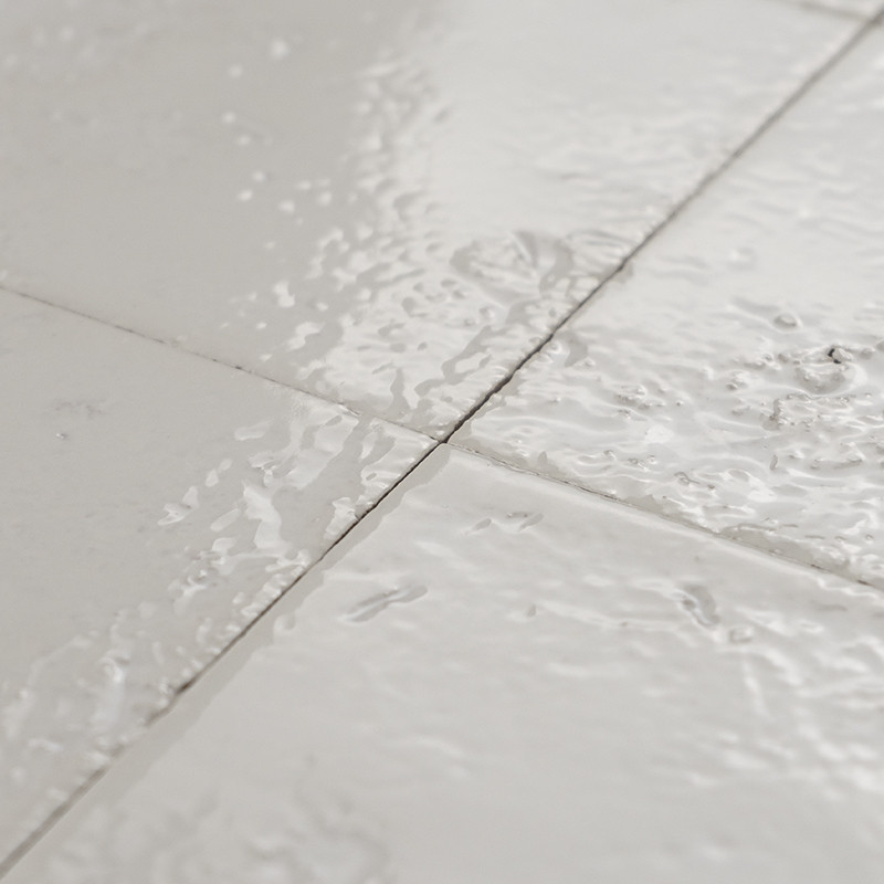 Bright white tile with visible joint and wet surface; context: adjacent tiles with reflections and wavy texture due to a film of water.
