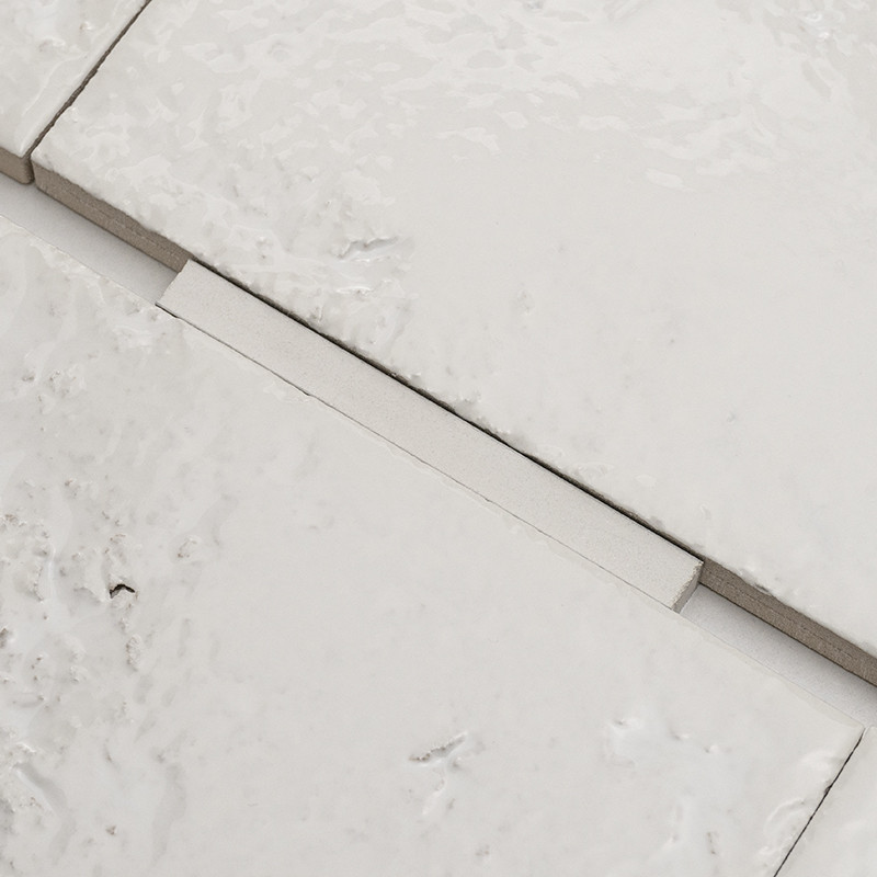 White ceramic tile, glazed surface with irregularities; fitted into gray grout joints, flanked by adjacent tiles and an elongated piece covering the joint.