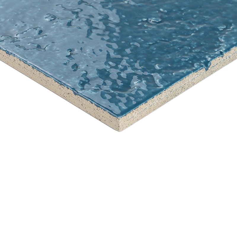 Carrelage bleu à surface brillante et ondulée, coin visible montrant couche support claire (dalle), bord biseauté sur fond blanc.