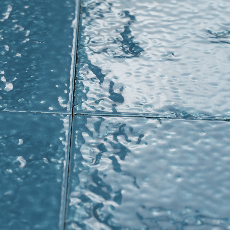 Blaue, glasierte quadratische Fliesen mit sichtbaren Fugen; glänzende, leicht gewellte, wasserähnlich reflektierende Oberfläche.