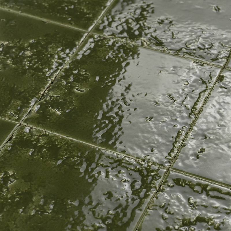 Bright green ceramic tile with irregular glaze; visible joints and rough texture, reflective surface with small accumulations, on a damp floor.