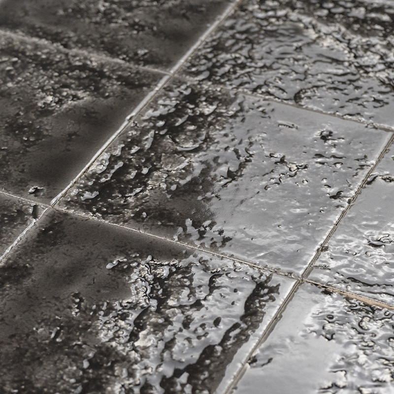 Glänzende, nasse schwarze Keramikfliesen mit sichtbaren Fugen und unregelmäßiger Oberfläche; Lichtreflexe und Wasserperlen betonen Struktur, aufgenommen aus kurzem Abstand.