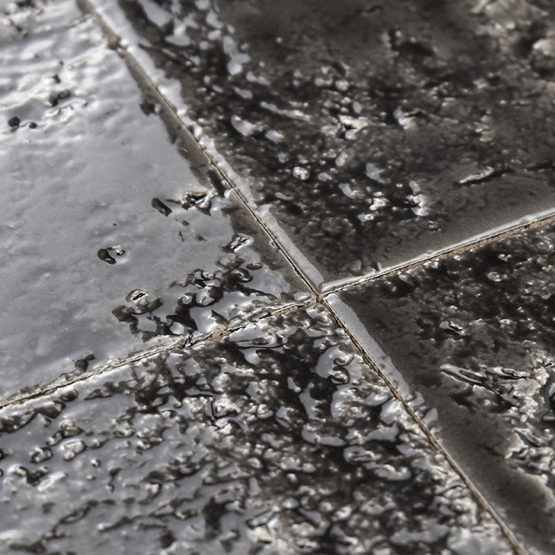 Carrelage quadrillé sombre et brillant, carreaux vernissés aux joints visibles, surface mouillée par des gouttelettes et reflets, aspect rugueux et irrégulier.
