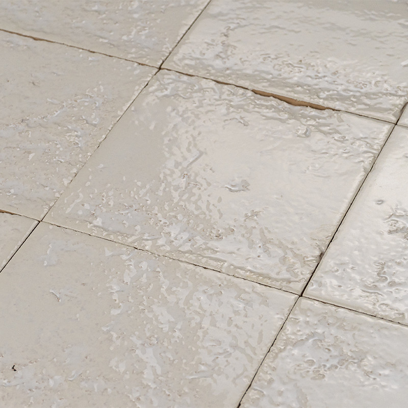 Azulejo cerâmico quadrado bege com acabamento vidrado irregular, rejuntes visíveis e pequena racha, superfície texturizada e reflexiva em piso interno.