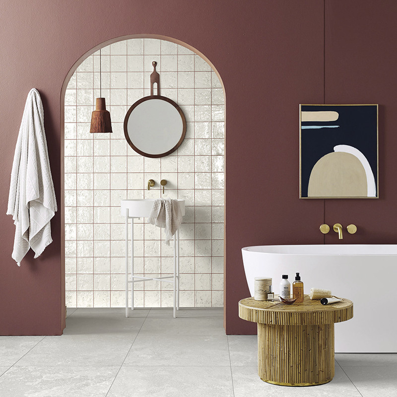 Weiße, glänzende quadratische Fliesen in bogenförmiger Nische; Kontext: Badezimmer mit bordeauxfarbenen Wänden, rundem Spiegel, kleinem Waschbecken, hängender Leuchte, freistehender Badewanne, Holzbeistelltisch und Handtuch.
