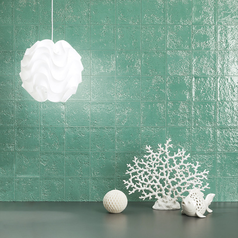 Grüne glänzende quadratische Fliesenwand; Vordergrund: dunkle Ablage mit links hängender weißer Lampe und rechts weißen Dekorobjekten (Korallenform, geriffelte Kugel, Vogelfigur).