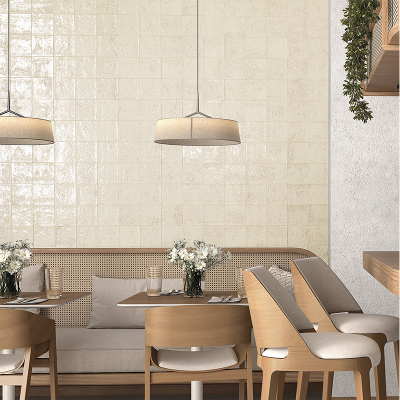 Azulejos cerâmicos creme brilhantes em parede; ambiente de café/cozinha com banco estofado rattan, mesa de madeira, cadeiras acolchoadas e pendentes; vasos com flores brancas.