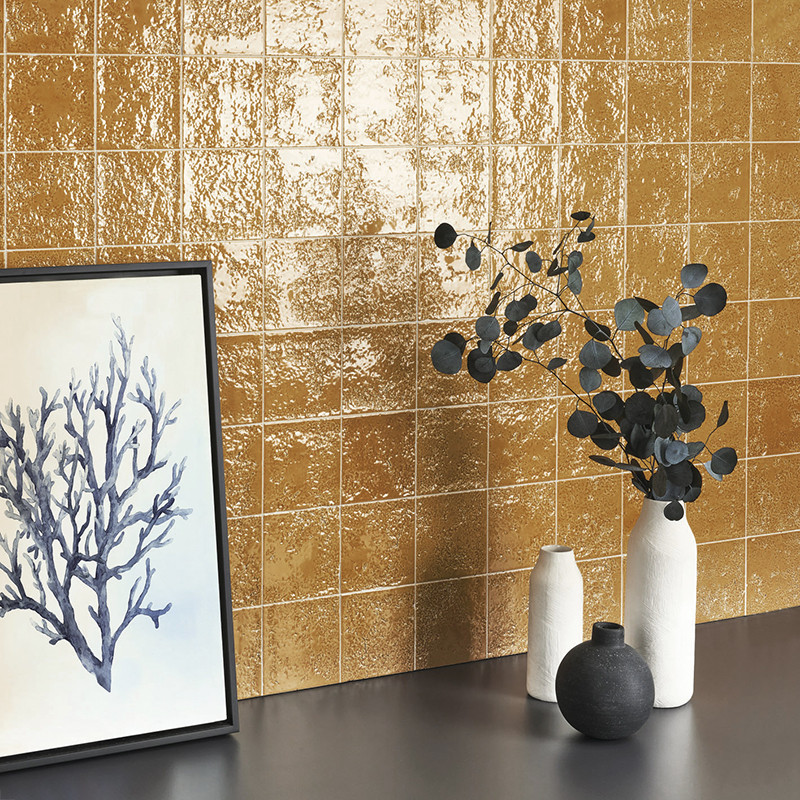Azulejos brillantes dorados, cuadrados y texturizados; sobre encimera oscura apoyan tres jarrones minimalistas y un cuadro con ilustración coralina, creando ambiente moderno y decorativo.