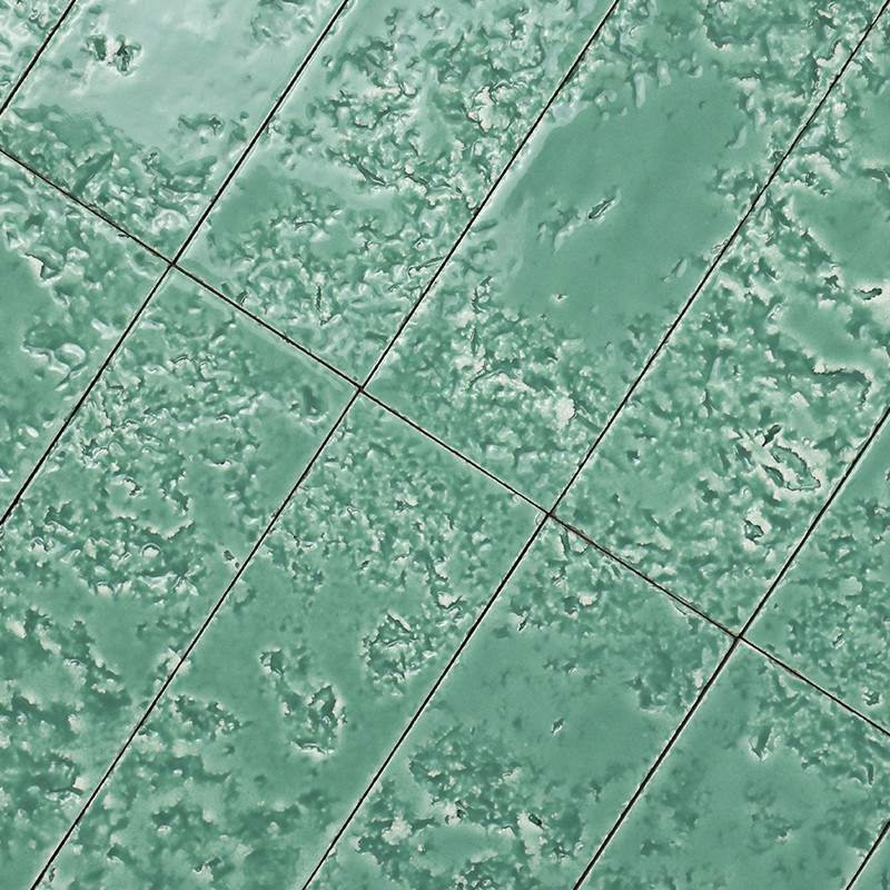 Groene keramische tegels met gemarmerde, licht reliëftextuur; diagonale donkere voeglijnen markeren individuele rechthoekige tegels; lijkt deel van vloer of wand.