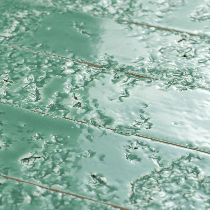 Geglazuurde groene tegels met ongelijkmatige bubbels en korrelige glazuurtextuur; diagonale voegen met lichtbruine kit, glanzend oppervlak en weerspiegelde lichtvlekken.