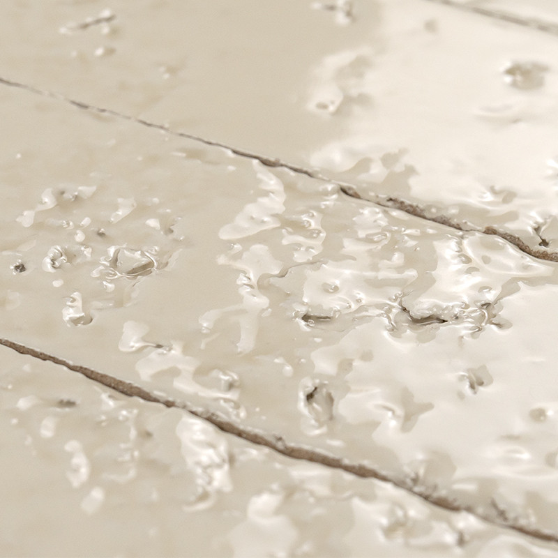 Azulejo claro con acabado brillante y gotas de agua dispersas; juntas oscuras y carriles que indican varias baldosas adyacentes, superficie reflectante y húmeda.