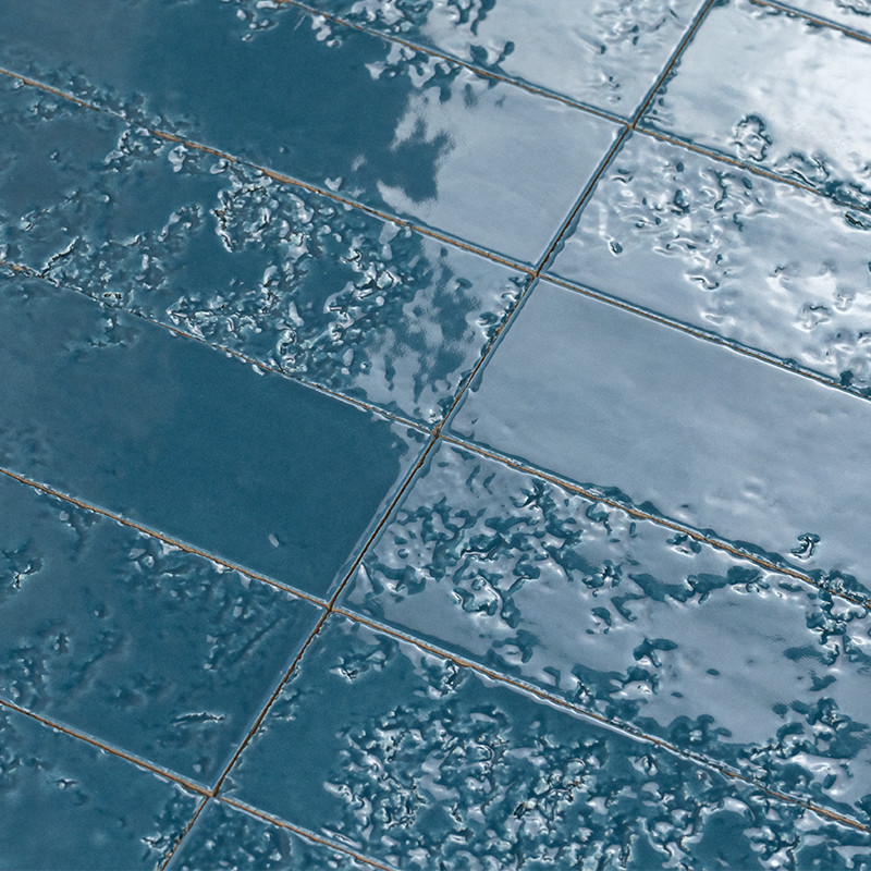 Azulejos cerámicos azul oscuro con esmalte irregular y textura burbujeada; juntas finas entre piezas; reflejos y sombras vegetales sobre la superficie.
