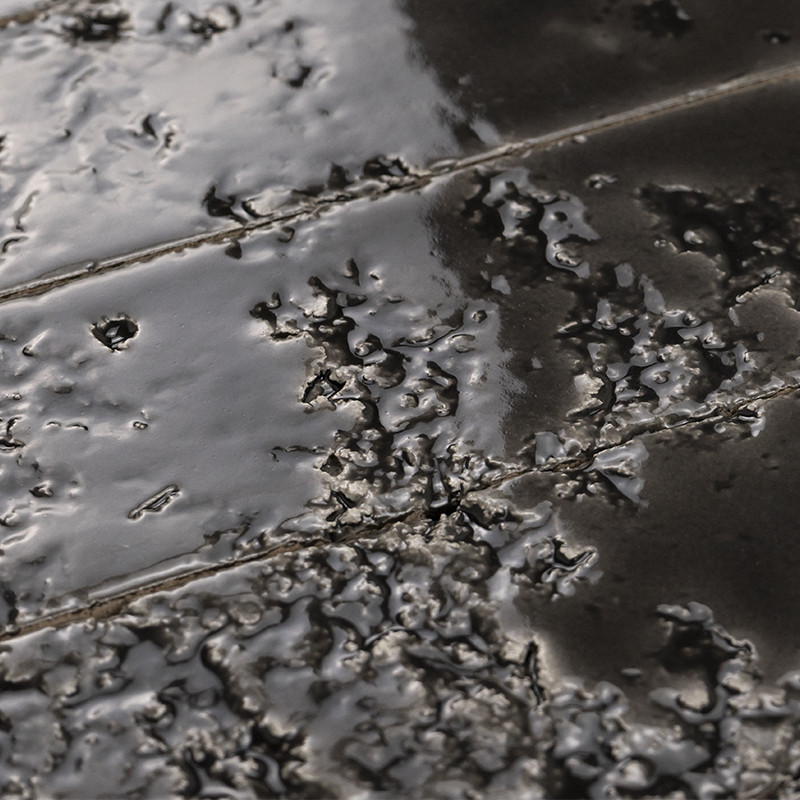 Azulejo esmaltado negro con superficie vidriada agrietada y textura burbujeante; juntas finas visibles; contexto cercano, reflejos brillantes sobre irregularidades.
