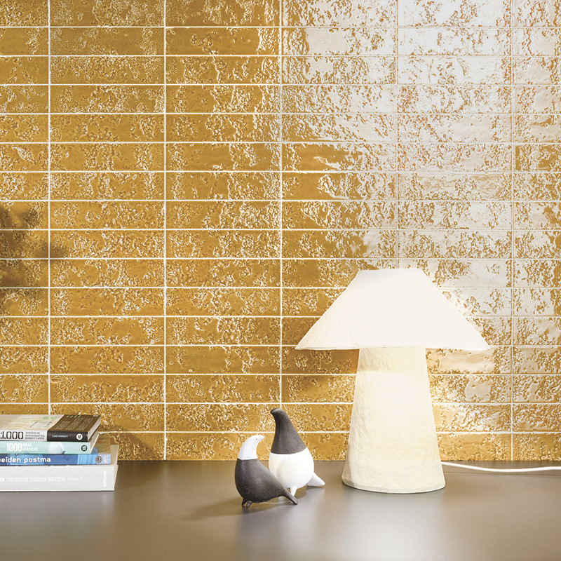 Goldfarbene, glänzende rechteckige Wandfliesen in Rastermuster; davor graue Arbeitsfläche mit cremefarbener Pilzlampe, zwei kleinen Vogelfiguren und einem Bücherstapel.