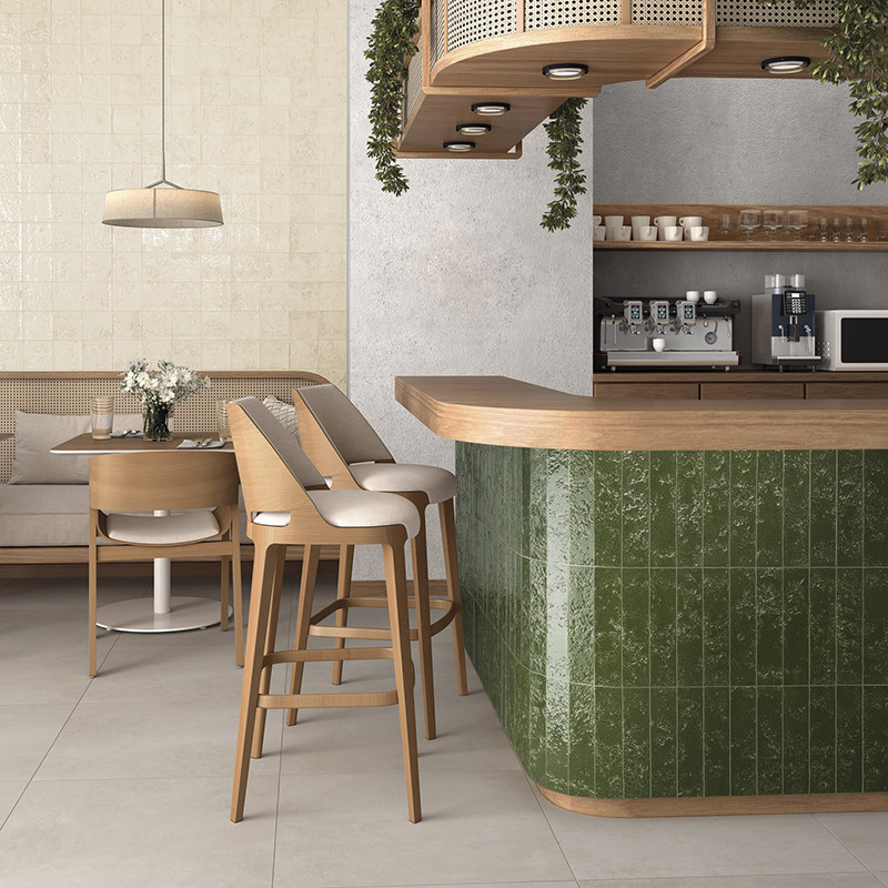 Textured green tile covers the curved bar; setting: modern cafe with upholstered wooden stools, table with flowers, shelves with coffee maker and beige wall with hanging lamp.
