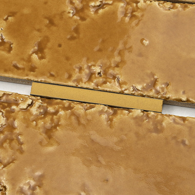 Rectangular ochre tile with textured surface and irregular shine; white joint between tiles and yellow rectangular spacer placed in the groove.
