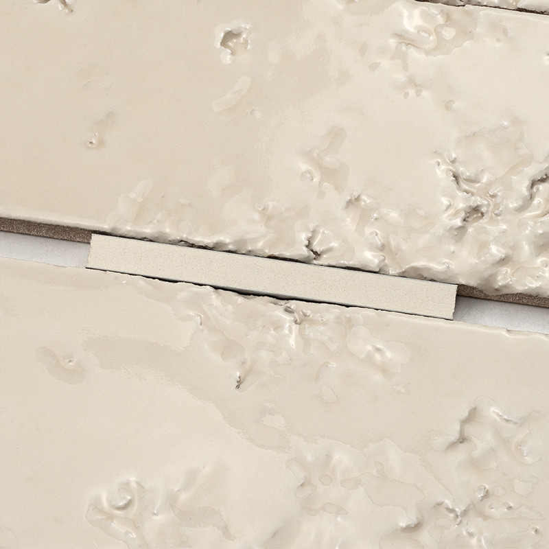 Azulejo creme texturizado com espaçador branco entre duas peças; superfície irregular com manchas e acúmulo de massa de rejunte no entorno.