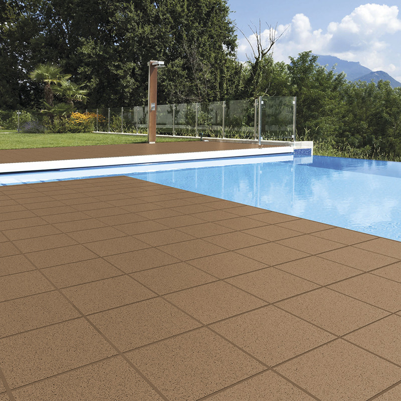Azulejos quadrados marrons em padrão de grade no deck, junto a piscina de água azul com borda branca, guarda-corpo de vidro, gramado, árvores e montanhas ao fundo.