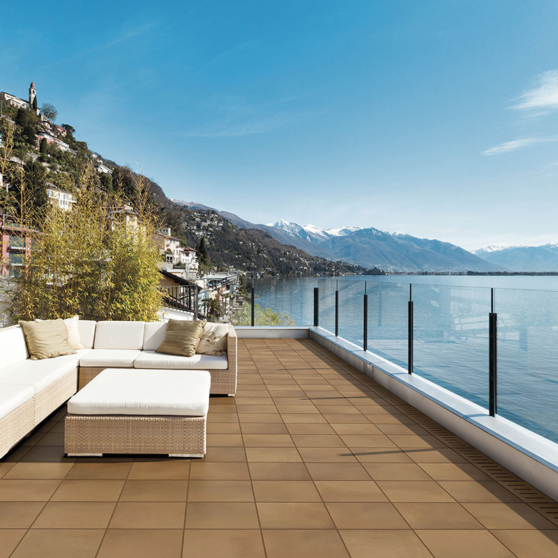 Vierkante bruine keramische terrastegels in rasterlay-out; moderne witte loungebank links, glazen balustrade rechts, uitzicht op bergachtig meer en heldere blauwe lucht.