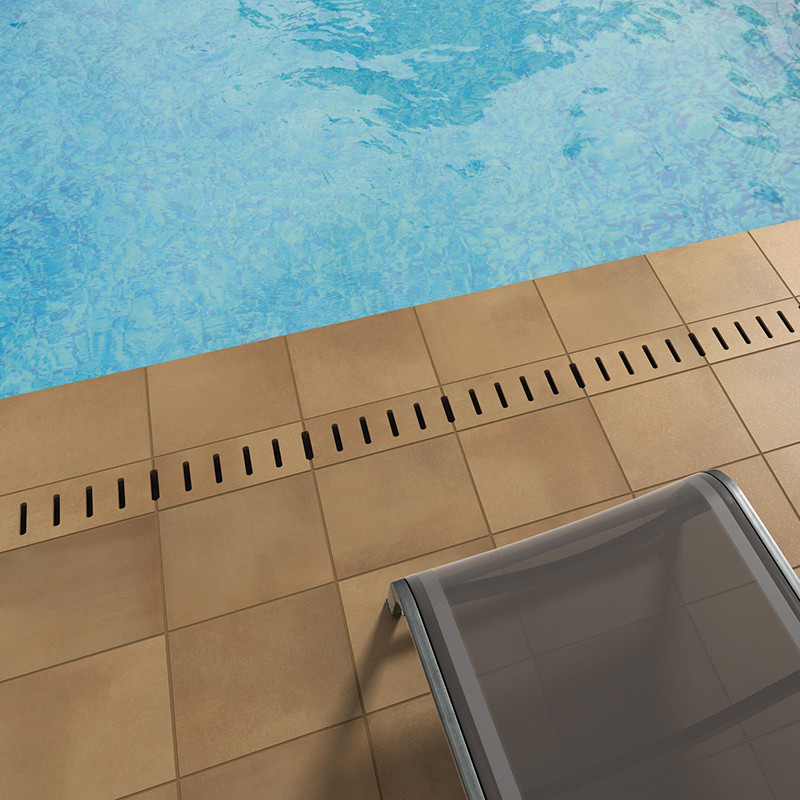 Beige paving tile next to a drainage groove; context: pool edge with blue water and a partially visible metal sun lounger.