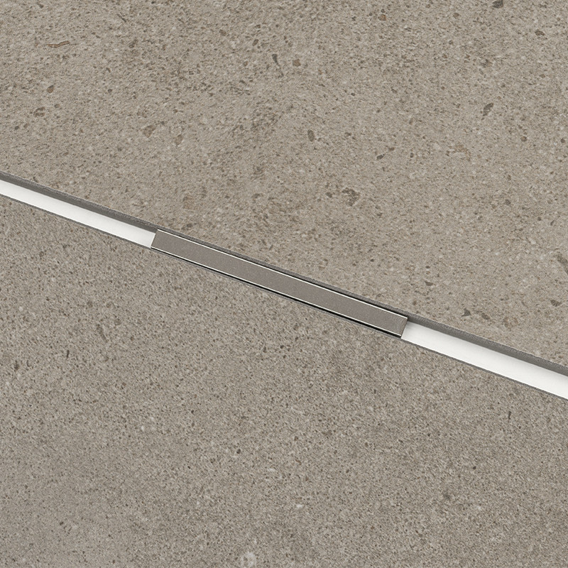 Rechteckige Sandstein-Fliese mit feiner Textur; diagonal eine schmale Fuge mit eingesetztem silbernem Metallprofil, Umgebung: ebene, graubraune Boden-/Wandfläche.