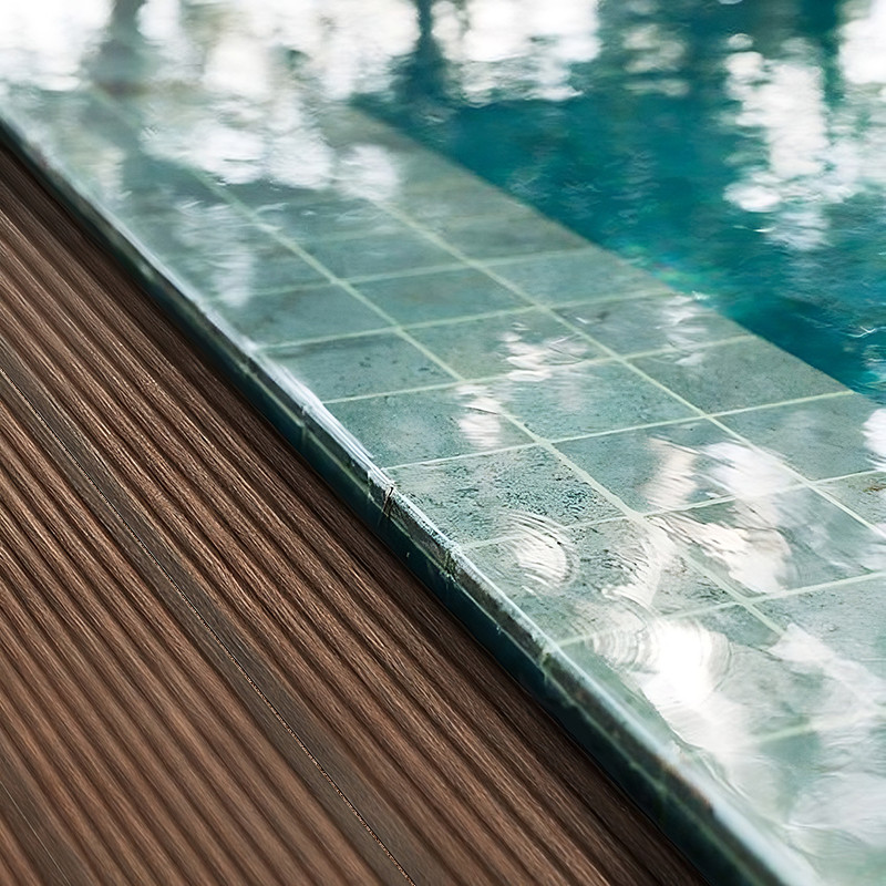 Carrelage vert pâle humide au bord d'une piscine, joints visibles et gouttelettes; contexte: terrasse en lames de bois rainurées à gauche, eau réfléchissante à droite.