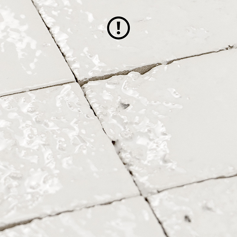 Carrelage blanc fissuré au centre, joints craquelés et résidus blanchâtres; angles ébréchés, surface brillante légèrement sale. Texte visible: "!"