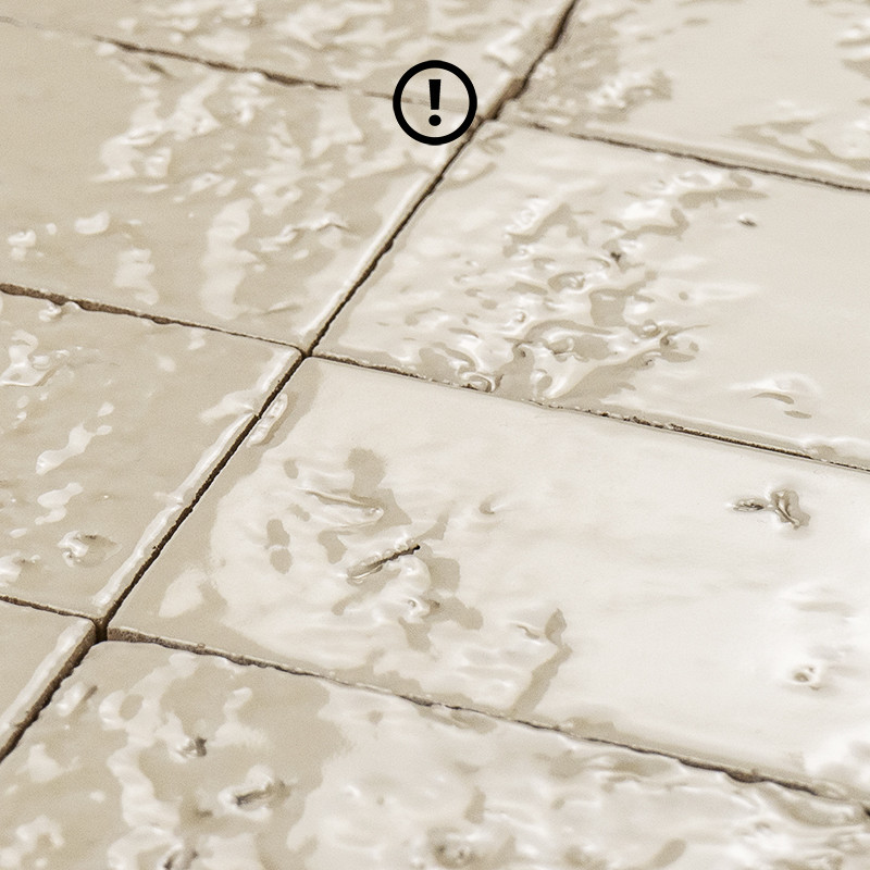 Azulejo cerâmico bege brilhante, superfície rugosa e irregular; rejuntes visíveis formando grade, close-up com um marcador circular contendo "!" sobreposto.