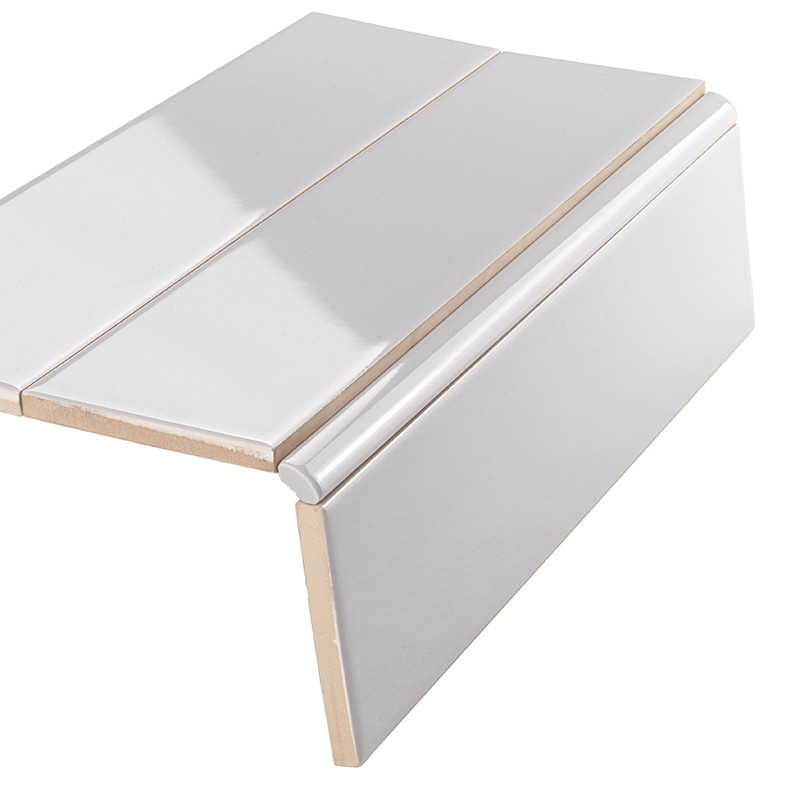 Witte glanzende keramische tegel met afgeronde afwerkstrip; gesneden randen tonen beige draaglaag, liggend tegen neutrale witte achtergrond.