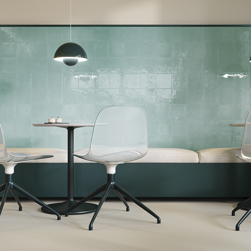 Türkis-glänzende quadratische Wandfliesen als Fokus; Kontext: moderne Café-Ecke mit gepolsterter Bank, runden Tischen, transparenten Stühlen und hängender schwarzer Pendelleuchte.