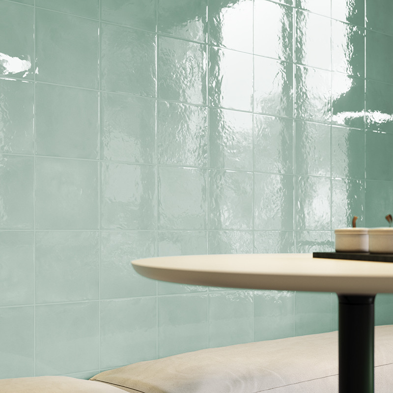 Azulejo cerámico esmaltado verde menta, superficie brillante y ondulada; pared detrás de una mesa redonda blanca con base negra y asiento acolchado claro, reflejos de luz natural.