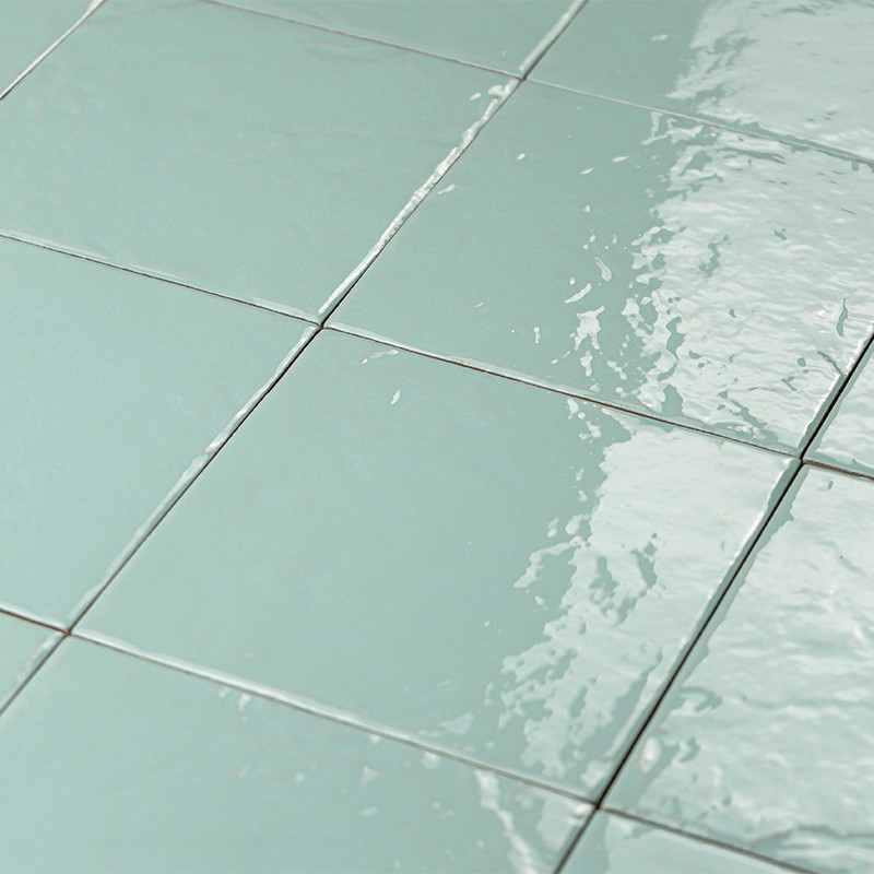 Azulejo esmaltado verde agua en cuadrícula, juntas claras; superficie brillante con reflejos y ligeras ondulaciones por agua, formando parte de un suelo o revestimiento cerámico.