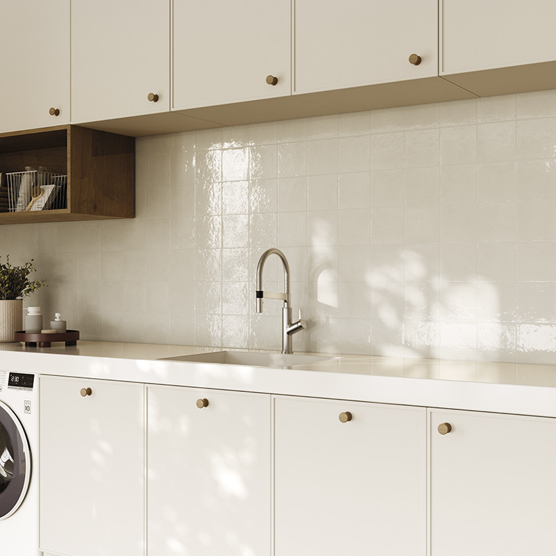 Azulejos brancos quadrados e brilhantes como backsplash; bancada branca com cuba e torneira inox, armários brancos com puxadores dourados, prateleira de madeira e máquina de lavar à esquerda.